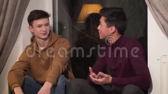 两个兴奋的男孩坐在窗台上说话年轻的亚洲男孩和白人朋友在室内交谈两个学院视频的预览图
