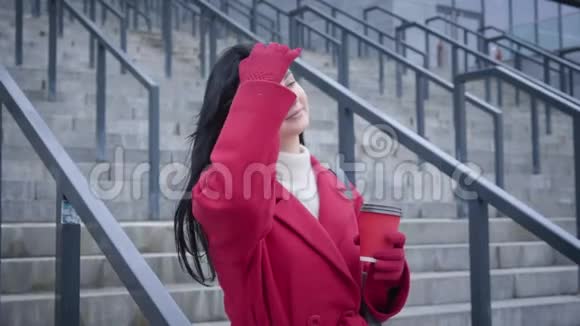 在城市里喝咖啡的迷人的白种人女士穿着红色外套和手套的肖像年轻开朗的女人视频的预览图
