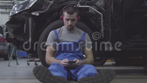疲惫的白种人男性汽车机械师靠在汽车车轮上听耳机里的音乐的肖像排气视频的预览图