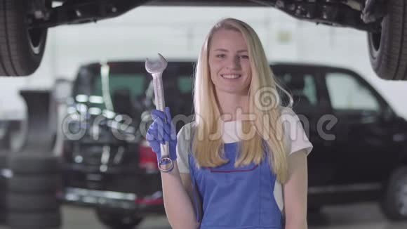 微笑的女汽车机械师穿着工作服拿着扳手伸出拇指金发白种人女人看着摄像机视频的预览图