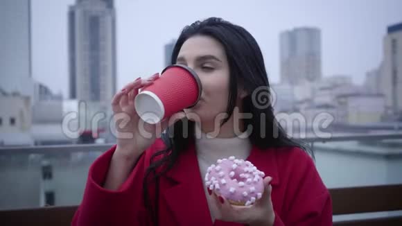可爱的白种人女孩绿眼睛黑发在户外喝咖啡吃香甜可口的甜甜圈视频的预览图