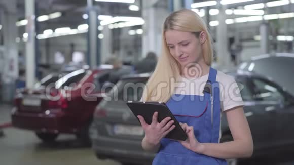 年轻微笑的白种人女人穿着工作服拿着平板电脑看着相机专业女汽车技师视频的预览图