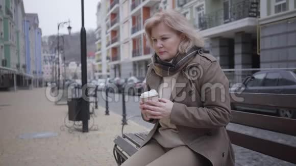 可悲的白人女人坐在户外的长凳上喝咖啡孤独优雅的退休者在城市街道上休息视频的预览图