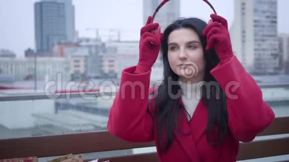 自信的白人女孩戴着红色外套和手套戴着耳机的肖像就像坐在城市里的小雨或视频的预览图