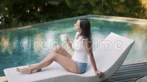 漂亮的赤脚女孩坐在游泳池旁的沙发上喝着美味的茶视频的预览图