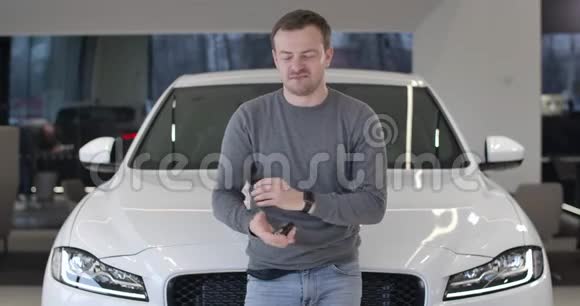 年轻的白人男子站在新的白色汽车和吹嘘汽车钥匙汽车幸福车主画像视频的预览图