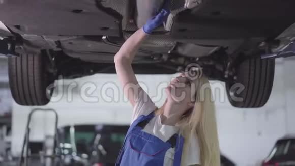 镜头在年轻的白种人汽车机械师周围移动检查汽车的底部在车里工作的严肃女人视频的预览图
