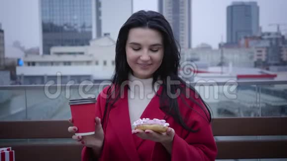 快乐的年轻白种人女人站在城市街道上拿着咖啡杯和甜甜圈看着相机微笑着肖像视频的预览图