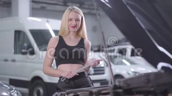 性感金发白种人女人的肖像站在旁边打开汽车引擎盖与可调扳手和微笑吸引力视频的预览图