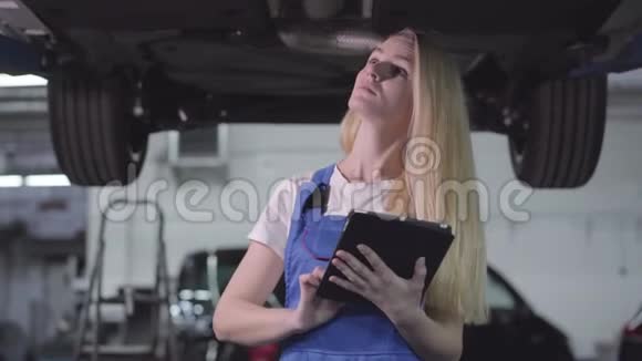 年轻美丽的白种人妇女穿着工作服站在破车下的平板电脑和注意失败的肖像视频的预览图
