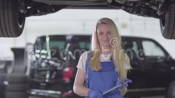 年轻的女汽车修理工穿着工衣站在修理厂的车下看着摄像机微笑着的画像视频的预览图
