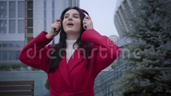 穿红色外套的活泼可爱的女孩走路和跳舞的肖像就像用耳机听音乐一样积极的高加索人视频的预览图