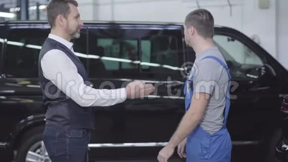 从侧面看白种人把车钥匙给汽车修理工成年商人与修理工交谈视频的预览图
