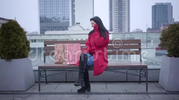 长镜头的开朗白种人女孩坐在小雨下在城市的长凳上并在电话年轻漂亮视频的预览图