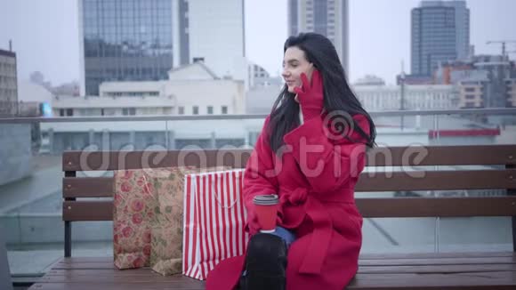 积极可爱的白人女性穿着红色外套坐在城市的长凳上拿着购物袋谈论着视频的预览图