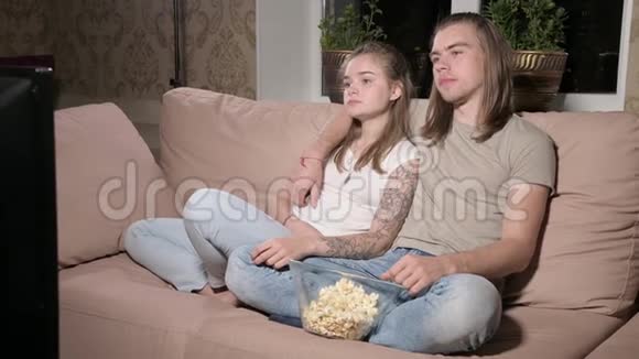 年轻可爱的夫妇坐在客厅里拥抱看电视吃爆米花视频的预览图