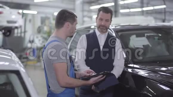 自信的白种人汽车机械师在平板电脑屏幕上向客户展示一些东西的肖像专业咨询视频的预览图