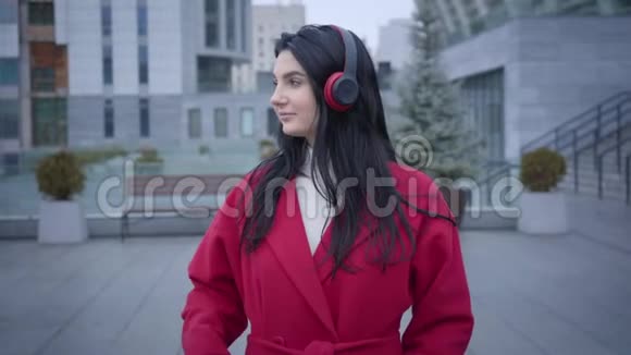 城市街道上年轻开朗的高加索女人戴着耳机跳舞的肖像穿红色外套的优雅女孩听着视频的预览图