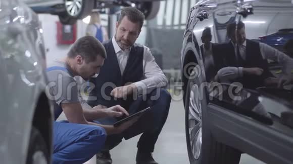 中镜头侧面的年轻白种人汽车机械师坐在客户旁边的汽车专业人士视频的预览图