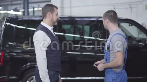侧视肖像年轻白种人汽车机械师持有汽车钥匙和与客户交谈工人和顾客视频的预览图