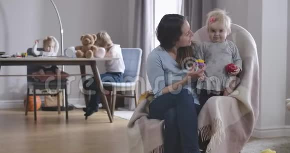 可爱的白种人女婴的肖像看着相机就像和妈妈坐在摇椅上一样她的姐姐和姐姐视频的预览图
