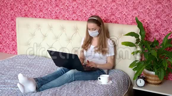 在家远程工作一个美丽的白种人女孩正在卧室里的笔记本电脑上工作床上戴着一个保护性的医用口罩和医生视频的预览图