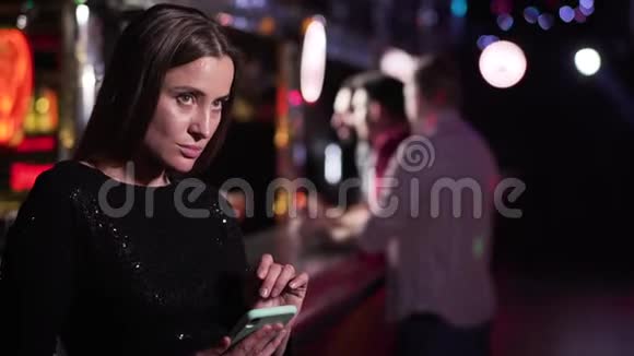 穿着黑色礼服站在夜总会酒吧柜台和使用智能手机的成年白人女性肖像视频的预览图