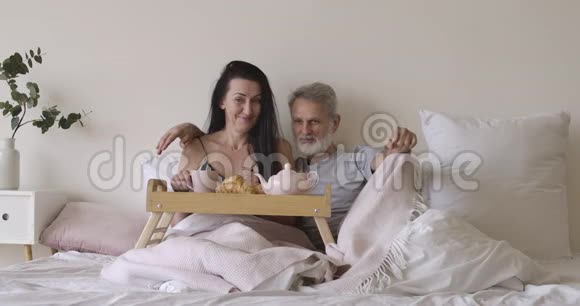 宽镜头肖像快乐的高加索人男女躺在卧室茶和羊角面包在床盘微笑视频的预览图