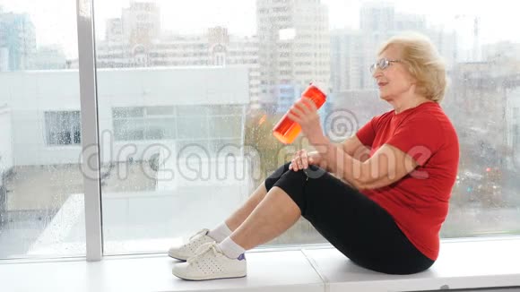 退休妇女享受晚年生活老白种人坐在窗台上从瓶子里喝水视频的预览图