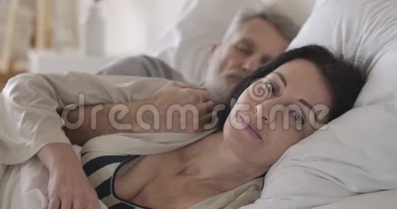 高加索黑发老妇人的特写镜头灰色眼睛表现出与熟睡的丈夫躺在床上的安静姿势视频的预览图