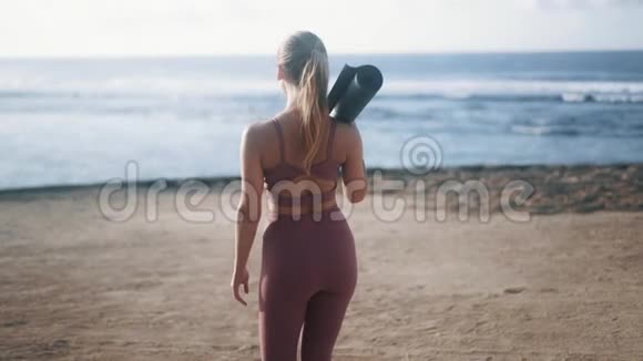 后视图穿着运动服带着体操垫的年轻女子去海滩视频的预览图