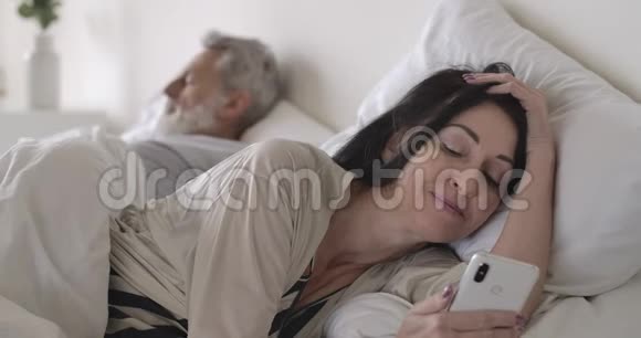 笑容满面的黑种人高级白种人女人使用社交媒体作为成熟的男人睡觉妻子早上躺在床上视频的预览图