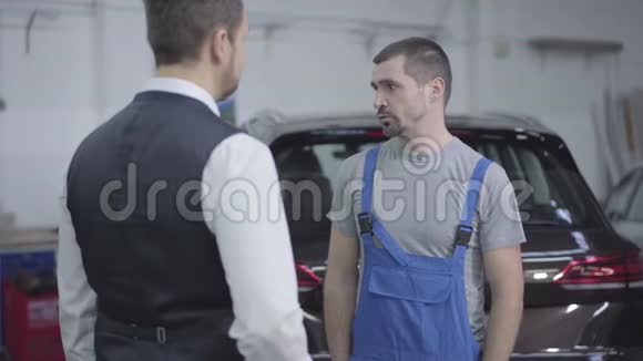 年轻的白种人穿着工作袍在修理厂拿着顾客的车钥匙摇着手汽车修理工开车视频的预览图