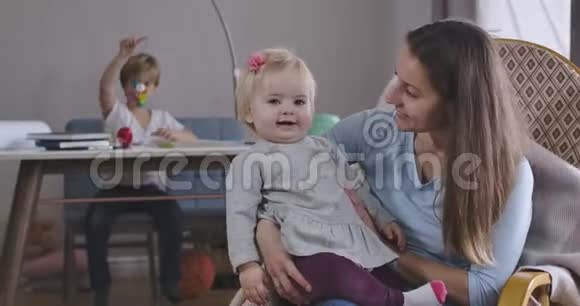 幸福的金发女孩和母亲坐在摇椅上微笑的肖像她哥哥在玩玩具视频的预览图