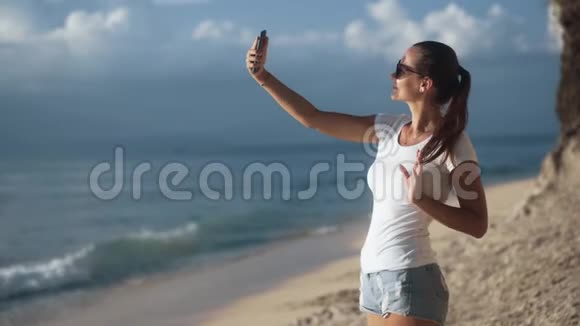 戴太阳镜的女人在日落时在靠近大海的智能手机上自拍视频的预览图