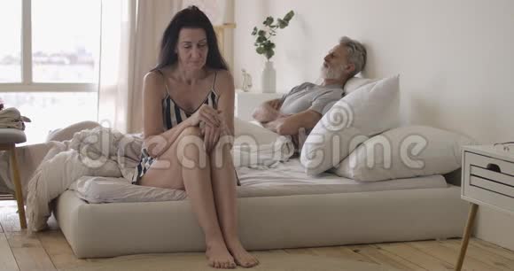一位体贴入微的白人高级妇女坐在床沿上双手抱着头丈夫躺在背景和背景上视频的预览图