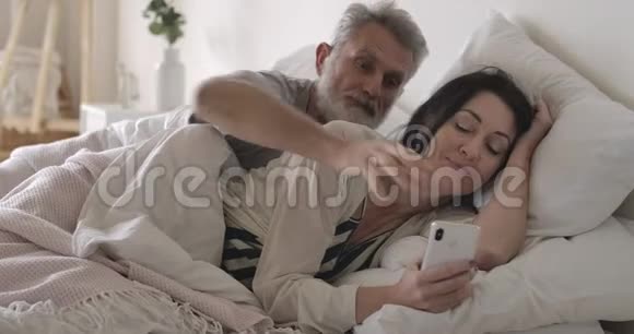 愤怒的高加索老人在床上拿走妻子智能手机丈夫愤怒地看着黑发女人在里面聊天视频的预览图
