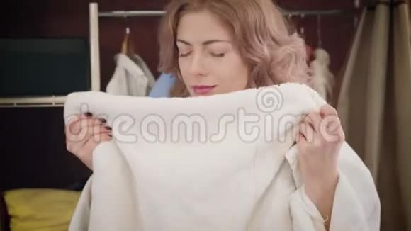 满意的年轻白种人女人的肖像穿着白色浴袍闻着并拥抱着干净的毛巾勤奋的家庭主妇视频的预览图
