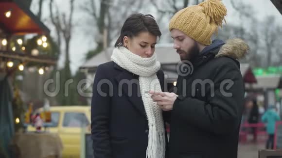 戴着黄色帽子戴着智能手机的白种人青年男子他的女朋友或妻子与他谈话时带有讽刺意味视频的预览图