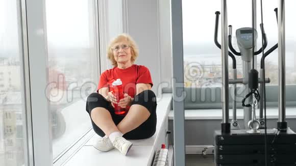 老年活跃的白种人妇女坐在窗台上在健身房训练后从瓶子里喝水退休妇女视频的预览图
