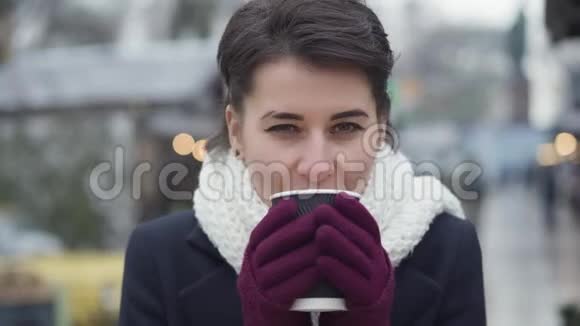 一个漂亮的高加索女人喝咖啡看镜头的特写镜头戴着白色冬巾的漂亮女孩视频的预览图