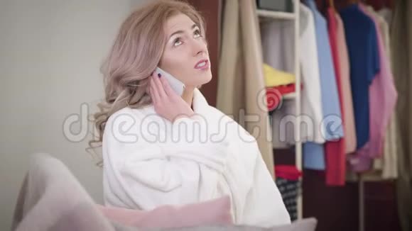 紧张的高加索女孩在智能手机上交谈的侧视美丽的年轻女子穿着白色浴袍坐在沙发上视频的预览图