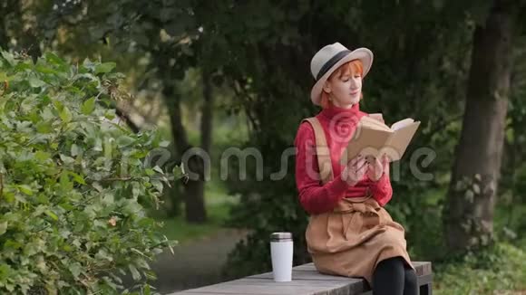 戴着帽子穿着毛衣的年轻红发女郎在长椅上看书喝着热咖啡视频的预览图