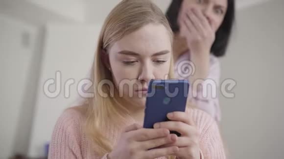 白种人模糊的女人用震惊的面部表情看着女孩肩膀上的智能手机屏幕的画像视频的预览图
