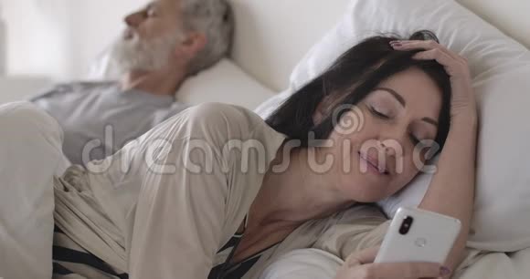 深色头发的白种人高级女性的特写照片她使用智能手机躺在床上作为丈夫睡在后台视频的预览图