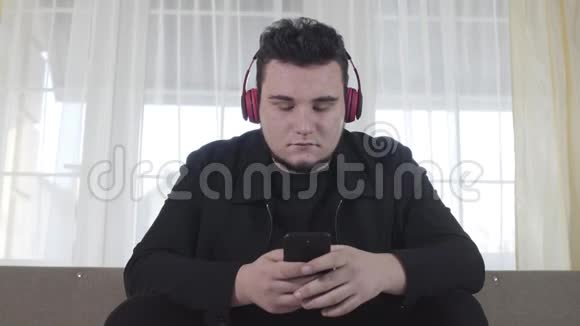 丰满的成年白种人戴着耳机坐在沙发上用智能手机的肖像听音乐的严肃男人视频的预览图