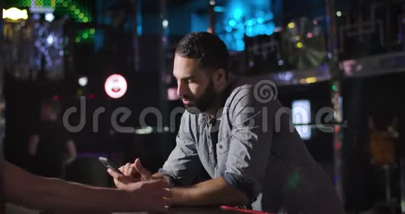 年轻的中东男子使用智能手机和饮酒酒吧柜台旁边的夜总会无聊的帅哥视频的预览图
