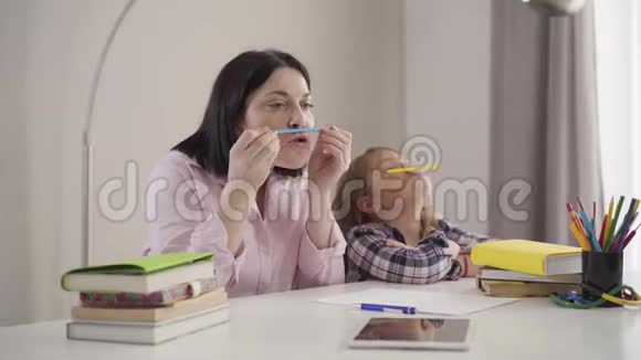 开朗的白种人女人和拿着铅笔和嘴唇的女学生的肖像妈妈和女儿玩得开心视频的预览图