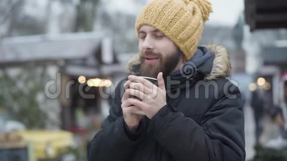 一幅年轻的白种人长胡子的肖像戴着滑稽的黄色帽子和冬天的外套在户外喝着热咖啡冷冻视频的预览图