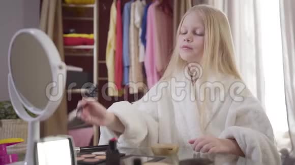 时尚的白人小女孩穿着白色浴袍坐在桌子旁带着化妆品和化妆金发可爱的孩子视频的预览图